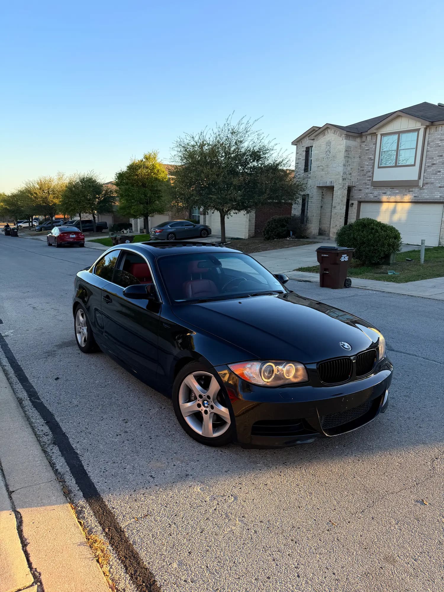 A black BMW 135i coupe, front three-quarter, on a residential street at golden hour with the paint washed and sealed.